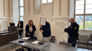 Ein Angeklagter am Gericht hält sich einen Ordner vor das Gesicht, neben ihm die Verteidigerin - Prozessbeginn am Landgericht Ellwangen: Ein islamischer Lehrer, Hodscha, wird beschuldigt, Jungen sexuell missbraucht zu haben.