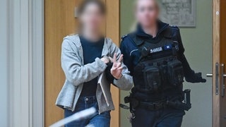 Ein Angeklagter wir von einer Justizwachtmeisterin in den Saal geführt.Am vierten Verhandlumgstag wegen des Angriffs auf ein Rüstungsunternehmen in Ulm hat ein erster Angeklagter ausgesagt.