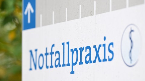 Auf einem Wegweiser an einer Straße vor einem Krankenhaus steht ein Hinweisschild auf eine Notfallpraxis. 