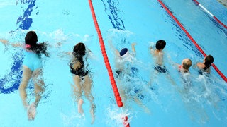 Schülerinnen und Schüler nehmen in einem Hallenbad am Schwimmunterricht teil. 