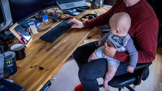 Ein junger Vater sitzt im Homeoffice mit seinem kleinen Sohn im Arbeitszimmer. In den Stellenanzeigen bieten immer mehr Arbeitgeber die Möglichkeit von Homeoffice an. Das ist das Ergebnis einer Bertelsmann Studie. 