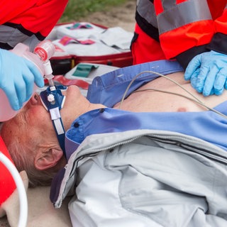 Ein Notarzt und ein Sanitäter versorgen einen Mann, der auf dem Boden liegt, mit Sauerstoff und führen eine Herzdruckmassage durch (gestellte Szene). 