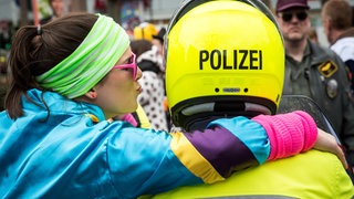 Ein Karnevalist von hinten mit einem Helm mit der Aufschrift "Polizist" wird umarmt von einer verkleideten Frau mit Stirnband und 80er-Jahre Jacke.