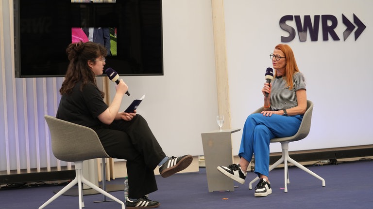 Moderatorin Mailin Engels (l.) hat SWR-Royal-Expertin Annelie Malun im Gläsernen Studio auf dem Maimarkt zu Gast. Sie sitzen im Gespräch auf der Bühne.