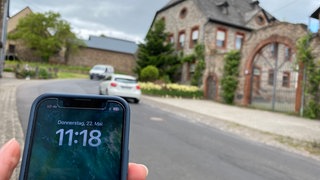 Ein Handy mit schlechtem Empfang, dahinter eine Dorfstraße. (Bürger und Bürgerinnen müssen in Münstermaifeld-Mörz (Kreis Mayen-Koblenz) oft auf der Suche nach Mobilfunkempfang gehen.)