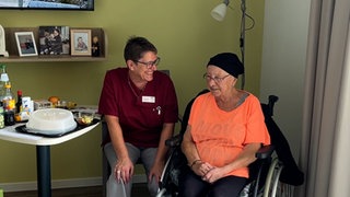 Schwester Wick und eine Patientin in einem Patientenzimmer. Sie lächeln einander an. 