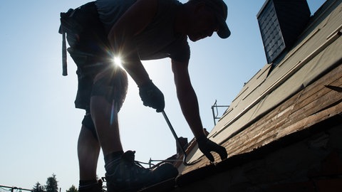 Ein Dachdecker steht bei gleißender Sonne auf dem Dach und arbeitet. Zunehmende Hitze trifft viele Branchen. Die Krankheitstage im Sommer nehmen auch in RLP stark zu.