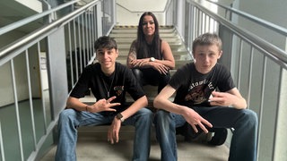 Gabriel (rechts), Justin (links) und Lehrerin Julia Molter sitzen auf einer Treppe im Schulgebäude