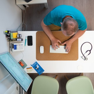 Ein Arzt sitzt an seinem Schreibtisch und tippt etwas auf seiner Tastatur ein. Eingeführt wurde sie bereits Ende April. Nun wird sie zur Pflicht: Ärztinnen und Ärzte müssen ab Oktober Diagnosen und Befunde in die elektronische Patientenakte (ePA) eintragen.