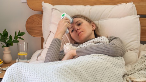 Eine kranke Frau liegt im Bett und hält sich ein Fieberthermometer an die Stirn. In Rheinland-Pfalz liegen die Grippezahlen noch auf niedrigem Niveau, die Corona-Zahlen aber steigen.