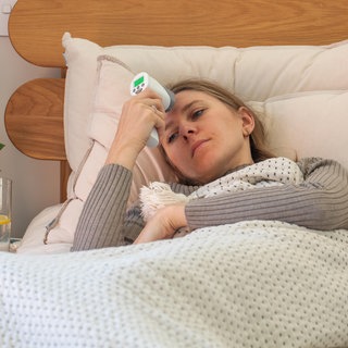 Eine kranke Frau liegt im Bett und hält sich ein Fieberthermometer an die Stirn. In Rheinland-Pfalz liegen die Grippezahlen noch auf niedrigem Niveau, die Corona-Zahlen aber steigen.
