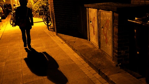 Eine Frau geht nachts im Licht einer Straßenlaterne auf einem Gehweg. 
