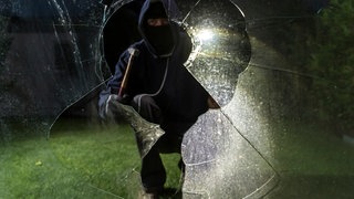 Ein maskierter Einbrecher leuchtet mit einer Taschenlampe in ein eingeschlagenes Fenster. Die Polizei hat eine mutmaßliche Einbrecherbande dingfest gemacht, die für mehr als 80 Einbrüche veranrwortlich sein soll.