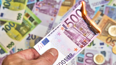 Eine Hand hält einen brennenden Geldschein. In Rheinland-Pfalz prüfen nun drei Staatsanwaltschaften, ob Steuergelder in Kommunen verschwendet wurden.
