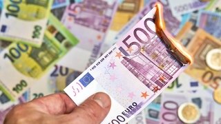 Eine Hand hält einen brennenden Geldschein. In Rheinland-Pfalz prüfen nun drei Staatsanwaltschaften, ob Steuergelder in Kommunen verschwendet wurden.