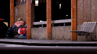 Eine obdachlose Frau sitzt in Decken eingehüllt an einem Bahnhof.