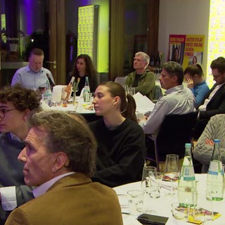 Besucher einer politischen Versammlung sitzen an runden Tischen und hören sich Vorträge an