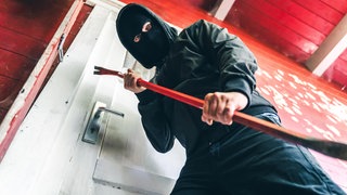 Maskierter Einbrecher steht vor einer Haustür mit Brecheisen (Symbolbild). Im vergangenen Jahr gab es weniger Staftaten in RLP.