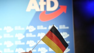 Die deutsche Flagge vor einem AfD-Hintergrund. Ein AfD-Mitarbeiter hat die Verfassungstreue-Überprüfung des rheinland-pfälzischen Landtags wegen seiner Vergangenheit in der "Revolte Rheinland" nicht bestanden und erhält kein Geld mehr aus der Staatskasse. 