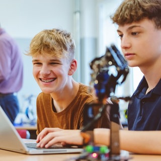 Zwei Schüler an einem Laptop und mit einem kleinen Roboter.