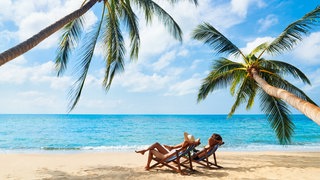 Eine Frau und ein Mann liegen auf Sonnenliegen an einem Strand unter Palmen.