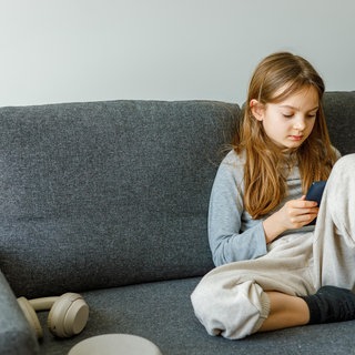 Ein Mädchen sitzt auf dem Sofa mit einem Smartphone. Digitale Endgeräte, Social Media und Künstliche Intelligenz bergen viele Gefahren, besonders für Kinder und Jugendliche (Symbolbild).