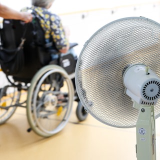 Eine Mitarbeiterin eines Seniorenheims schiebt eine Bewohnerin über den Flur, während ein Ventilator einen kühlen Luftzug erzeugt.