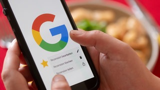 Hände halten vor einem gedeckten Esstisch ein Smartphone mit dem Google-Logo und dem Menüpunkt eine Rezension zu bearbeiten, zu löschen oder zu teilen.