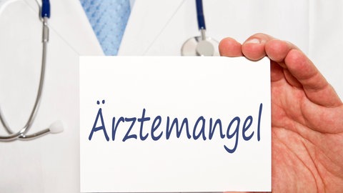 Ein Arzt mit einem Schild, auf dem Ärztemangel steht (Symbolbild)
