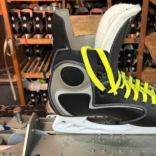 Eislaufschuh in einem Kufen-Schleifgerät, im Hintergrund mehrere Eislaufschuhe in Holzregalen