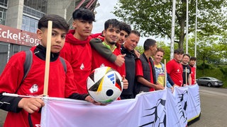 Schüler der Nina-Pfaff-Realschule-plus und Organisator Marian Prill stehen vor dem Horst-Eckel-Tor am Fritz-Walter-Stadion auf dem Betzenberg. Hier in Kaiserslautern wird angestoßen für die besondere Fan-Aktion zur Fußball-EM.