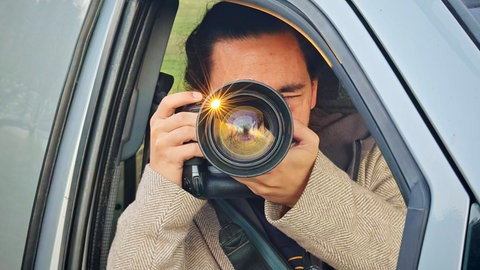 Ein Mann sitzt in einem Auto und hält eine Kamera mit großem Objektiv vor das Gesicht. (Symbolbild)