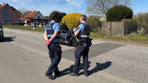 Zwei Polizeibeamte beobachten am Straßenrand in Mehlingen bei der Grundschule den durchfahrenden Verkehr. Gerade passiert ein schwarz gekleideter Motorradfahrer die Straße. 