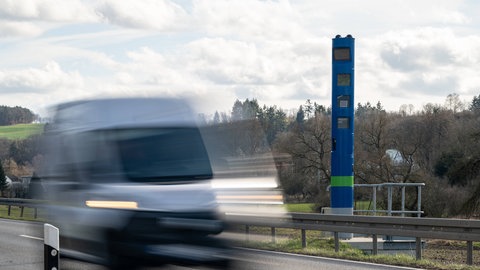 Ein Bild zeigt eine blaue Stehle zur Erfassung der Lkw-Maut auf einer Landstraße, ein Transporter fährt daran vorbei. (IMAGO)