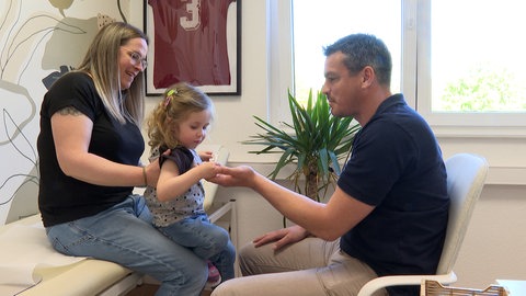 Kinderarzt Peter Follmann zaubert in seiner Praxis in Winnweiler am Donnersberg ein Gummibärchen hervor. Das gefällt der zweijährigen Anna. Sie sitzt auf dem Schoß von Mama Christine Schneider. 