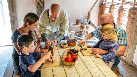 Eine Familie sitzt im Keltendorf in Steinbach am Tisch. Sie arbeiten gemeinsam mit Hammer und Nagel.