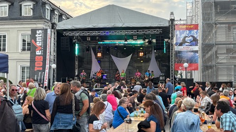 In Zweibrücken wurde am Wochenende das Stadtfest gefeiert. Auf dem Herzogplatz war viel los. Viele Leute sitzen auf Bierbänken vor der großen Bühne, auf der eine Band spielt.