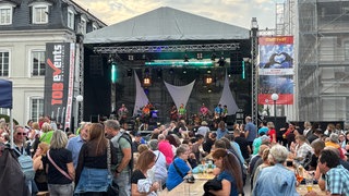 In Zweibrücken wurde am Wochenende das Stadtfest gefeiert. Auf dem Herzogplatz war viel los. Viele Leute sitzen auf Bierbänken vor der großen Bühne, auf der eine Band spielt.
