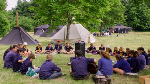 Die Jugendzeltplätze in der Pfalz haben trotz des Wetters eine gute Auslastung, so auch der Jugendzeltplatz Hauenstein. (Pfadfinder-Gruppe sitzt in einem Kreis auf einem Zeltplatz.)