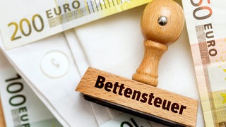 Stempel mit Aufschrift Bettensteuer und Geldscheine auf einem Bettlaken.