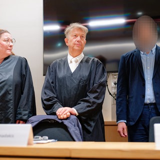 Der Angeklagte (rechts) steht im Verhandlungssaal neben seinen Rechtsanwälten