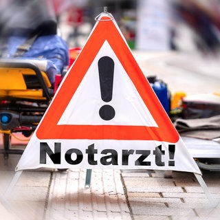 Die Fotomontage zeigt ein Warndreieck mit der Aufschrift "Notarzt" vor einer verschwommenen Rettungseinsatz: Bei einem Herz-Kreislauf-Stillstand dauert es im Bereich des Rettungsdienstbereichs Montabaur oft länger als vorgeschrieben, bis Hilfe kommt.