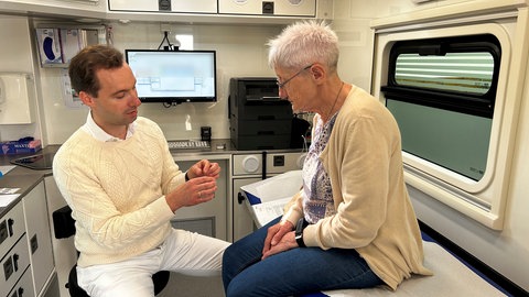 Ein Arzt spricht in einer rollenden Arztpraxis mit einer älteren Frau: Für viele Menschen in Herdorf ist die rollende Praxis eine Erleichterung, weil der Hausarzt zu ihnen kommt und nicht umgekehrt. 