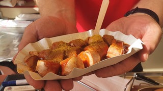 Eine Frau hält eine Currywurst in einer Schale in einem Imbisswagen.