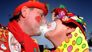 Zwei Narren geben sich bei einem Fastnachtsumzug bei strahlenden Sonnenschein ein Küsschen. In Rheinland-Pfalz wird am Rosenmontag schönes Wetter mit viel Sonne.