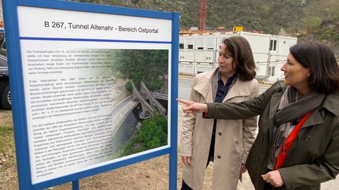 In Altenahr schauen sich Cornelia Weigand (links), die Landrätin des Kreises Ahrweiler, und Wirtschaftsministerin Daniela Schmitt (rechts, FDP) eine Infotafel an. Dort wurde der Tunnel der B267 nach der Flutkatastrophe saniert.