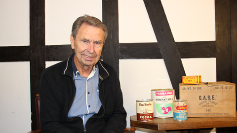 Günter Weckbecker aus Moselkern sitzt an einem Tisch mit einem CARE-Paket, davor stehen alte Dosen mit Milchpulver und Dosenfleisch. 