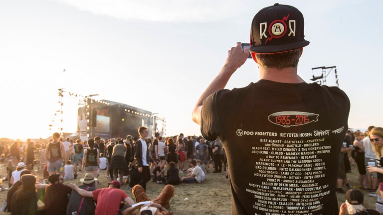 Ein Festivalbesucher mit einem Rock am Ring-Shirt steht vor der Bühne und macht ein Foto. Das aktuelle Festivalshirt ist für viele Fans fast schon ein Muss.