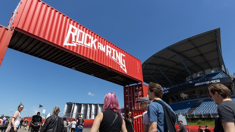 Besucher gehen unter einer roten Containerbrücke mit der Aufschrift "Rock am Ring" das Festivalgelände: Rock am Ring gibt es 2025 seit 40 Jahren am Nürburgring. 