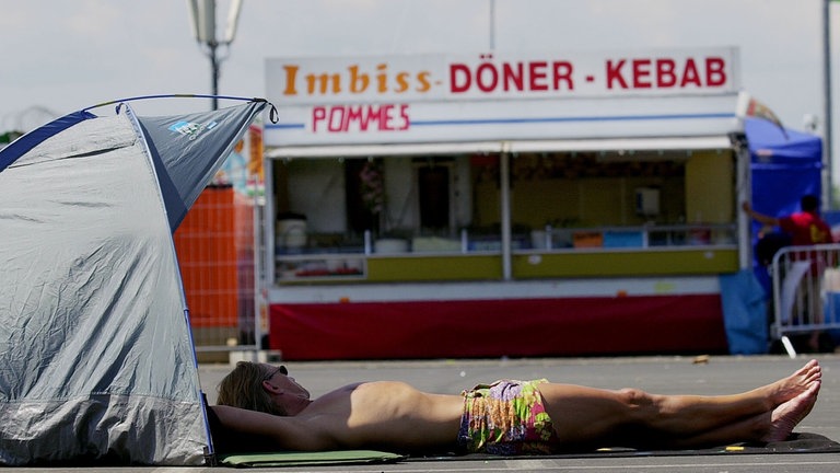 Bei Rock am Ring 2003 liegt ein halbnackter Fan auf dem Asphalt vor einer Dönerbude in der Sonne und macht eine Pause.